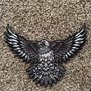May include: Embroidered patch featuring a detailed eagle with outstretched wings. The patch is primarily black and gray, with white accents on the feathers. The eagle is centered, showcasing its sharp beak and focused gaze. This patch is suitable for jackets, bags, or other fabric items.
