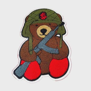 May include: Embroidered patch featuring a brown teddy bear wearing a green military helmet with a red emblem and holding a blue rifle. The bear has red feet and a black nose and eyes. The patch has a white border.