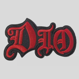 May include: Embroidered patch featuring the word "DIO" in a gothic font. The letters are red with a black border and backing. The patch is designed for application on clothing or accessories.