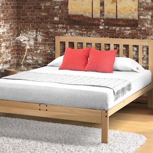 May include: A light-colored wooden bed frame with a headboard featuring vertical slats. The bed is made up with a white mattress, two red pillows, and a decorative throw. The bed sits on a white rug in front of a brick wall.