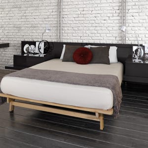 May include: A modern bedroom with a light wood bed frame, a white mattress, and a gray throw blanket. The bed has a black headboard with black nightstands on either side. Decorative items and lamps are on the nightstands. The room has a brick wall.