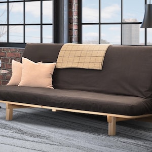 May include: A brown futon sofa with light wood legs and a matching brown cushion. Two peach-colored pillows and a tan blanket rest on the sofa. The sofa sits on a gray rug in front of a brick wall.