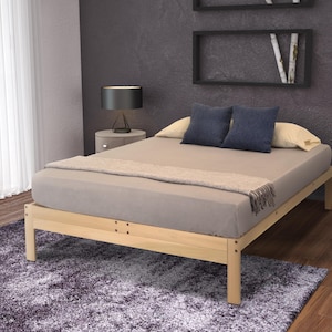 May include: A light-colored wooden bed frame with a mattress, pillows, and a blanket. The bed frame has a simple, rectangular design. The bed is covered with a gray sheet and a beige blanket with fringe. Two blue pillows and a yellow pillow are on the bed.