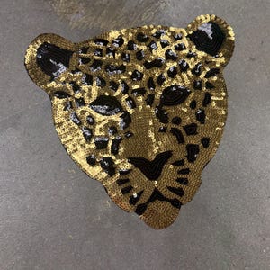 May include: A sequined leopard head patch in gold, black, and white. The patch features a detailed design with gold sequins forming the base, black sequins outlining the features, and white sequins adding highlights. The leopard's face is centered, with ears at the top.