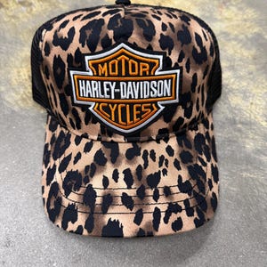 May include: A trucker cap featuring a tan and black leopard print pattern on the front panels and brim. The centre of the cap displays an embroidered Harley-Davidson Motor Cycles shield logo in orange, white, and black thread with black mesh sides.