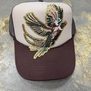 Puede incluir: Gorra de camionero marrón y beige con un diseño detallado de pájaro con lentejuelas. El pájaro presenta lentejuelas verdes, doradas y marrones, con un ojo rojo. La gorra tiene una visera marrón y una parte trasera de malla.