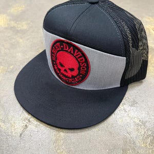 Op de afbeelding: Een zwart-grijze truckerpet met een platte klep en een achterkant van mesh. Het voorpaneel is voorzien van een ronde patch met een rode schedelafbeelding en de tekst Harley-Davidson Motorcycles in zwarte letters langs de rand geborduurd.