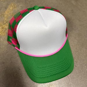 May include: A trucker hat featuring a solid white foam front panel, a green brim, and a bright pink braided cord trim. The side and back panels are constructed from a green and pink checkered mesh fabric, topped with a small green button.