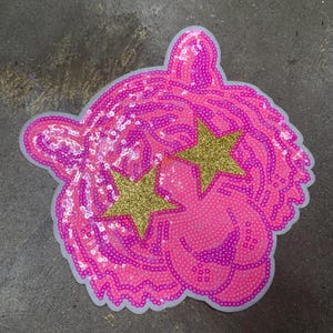 May include: A pink sequined tiger head patch with gold glitter star eyes. The patch has a white border and is likely for clothing or accessories. The design is bold and eye-catching, suitable for adding flair to garments.