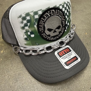 Può includere: Un cappellino trucker grigio e bianco con una toppa circolare nera raffigurante un teschio e la scritta Harley-Davidson Motorcycles. Il cappellino presenta una catena color argento sulla visiera e un adesivo con la scritta OTTO World Famous Foam Front.