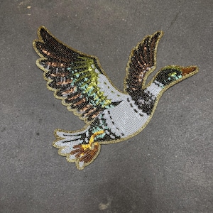 May include: A decorative duck patch with a gold outline, wings spread in flight. The patch is embellished with sequins in shades of brown, green, white, and black. The duck's beak is a light brown color.