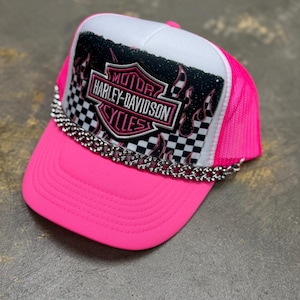 May include: A bright pink trucker cap with a white top panel. It features a Harley-Davidson Cycles logo with a chequered background and a silver chain along the brim. The logo is in black and pink.