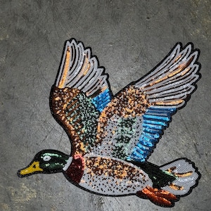 May include: A sequined patch of a duck in flight. The duck has a green head, brown and white body, and wings with blue, orange, and silver sequins. The patch is outlined in black.
