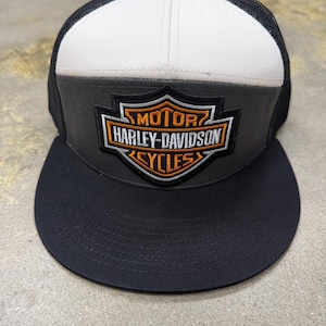 Op de afbeelding: Een zwart-witte truckerpet met een donkergrijs voorpaneel voorzien van een geborduurd Harley-Davidson Motor Cycles schildlogo in oranje, wit en zwart. De pet heeft een effen zwarte platte klep en een ademend zwart mesh achterpaneel.