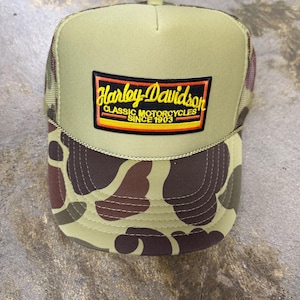 May include: A tan and brown camouflage trucker hat featuring a rectangular patch on the front. The patch displays the text Harley-Davidson in yellow script, with Classic Motorcycles Since 1903 in white text, bordered by orange and black lines.