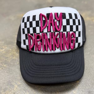 Puede incluir: Gorra de camionero en blanco y negro con un patrón de cuadros en el panel frontal. La gorra presenta el texto bordado "DAY DRINKING" en rosa. La visera es negra y la parte trasera de malla es negra.