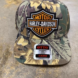 May include: A camouflage print baseball cap featuring a black and orange Harley-Davidson Motor Cycles shield patch on the front. The cap includes an Otto brand sticker on the brim with text reading Premium Fabric, Classic Fit, OSFM, and Camo.