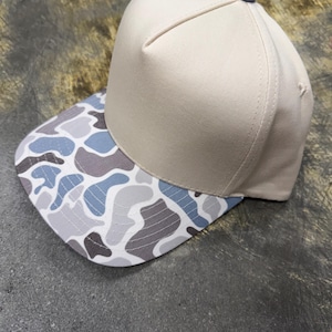 Op de afbeelding: Een baseballpet met een effen beige kroon en een platte klep met een meerkleurig camouflagepatroon in tinten lichtblauw, donkergrijs en wit. De pet heeft een kleine blauwe knoop in het midden bovenaan waar de panelen samenkomen.