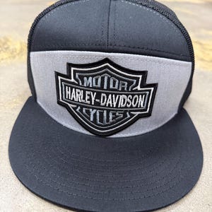 May include: A black and grey baseball cap featuring a mesh back and a flat brim. The front panel displays a prominent black and white embroidered shield-shaped patch with the text MOTOR HARLEY-DAVIDSON CYCLES centered within the design.