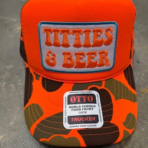 Pode incluir: Um boné trucker laranja brilhante com um emblema retangular com o texto TITTIES & BEER bordado em laranja. A aba apresenta um padrão de camuflagem laranja e castanho escuro. Um autocolante branco do produto está fixado na aba frontal.