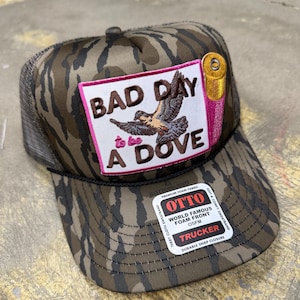 Könnte beinhalten: Eine Camouflage-Trucker-Cap mit braun-grünem Muster, die einen rechteckigen Aufnäher mit dem Text "BAD DAY to be A DOVE" sowie eine gestickte Taube und eine Schrotpatrone zeigt. Die Kappe hat eine schwarze Kordel am Schirm und einen Otto-Aufkleber.
