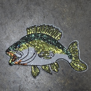 May include: A sequined fish applique in shades of green, gold, and silver. The fish has an open mouth with orange accents and is set against a gray background. The design features detailed sequin work, creating a textured and shimmering effect.