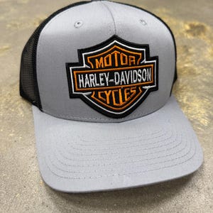 Puede incluir: Una gorra de béisbol gris con parte trasera de malla negra y panel frontal estructurado. El frente presenta un logotipo de escudo Harley-Davidson Motor Cycles bordado en hilo naranja, blanco y negro. La gorra tiene una visera plana y un botón.