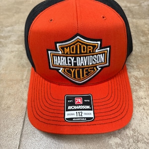 Może przedstawiać: Pomarańczowo-czarna czapka typu trucker z wyhaftowanym z przodu logo tarczy Harley-Davidson Motor Cycles. Czarno-biała naklejka na daszku zawiera napis Est. 1970 Richardson Original 112 Trucker Adjustable.
