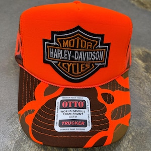 May include: A bright neon orange and brown camouflage trucker hat featuring a prominent Harley-Davidson Motor Cycles shield patch on the front foam panel. A rectangular sticker on the brim displays the text OTTO, PREMIUM FOAM FABRIC, and TRUCKER.