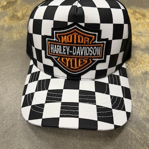 May include: Black and white checkered baseball cap with a mesh back. The cap features an embroidered Harley-Davidson logo in orange and black on the front. The brim is also checkered.