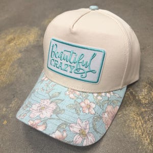 Pode incluir: Um boné de beisebol cor de creme com um emblema retangular na frente com o texto "Beautiful CRAZY" bordado em azul-petróleo. A aba apresenta um fundo azul-claro com um padrão repetitivo de flores cor-de-rosa e folhas verdes.