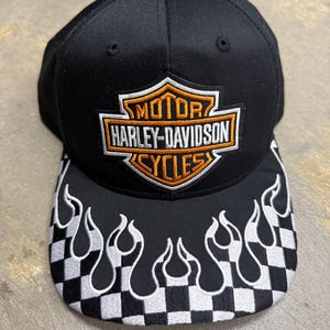 May include: A black baseball cap featuring an embroidered Harley-Davidson Motor Cycles shield logo in orange, white, and black on the front crown. The brim displays a white and black checkered pattern overlaid with white flame embroidery.
