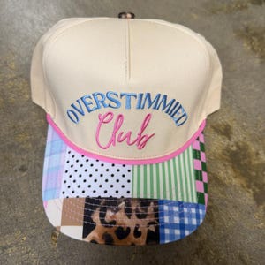 Op de afbeelding: Een crèmekleurige baseballpet met de geborduurde tekst OVERSTIMMIED in blauw en Club in roze. De klep heeft een veelzijdig patroon met zwarte stippen, groene strepen, een bruine luipaardprint en een blauw-wit ruitjespatroon.