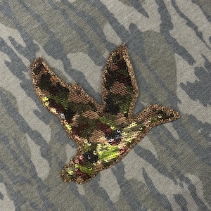 May include: A fabric garment featuring a camouflage pattern in shades of gray and green. Centered on the fabric is a duck-shaped appliqué covered in shimmering sequins that display a mix of green, brown, and black camouflage colors.