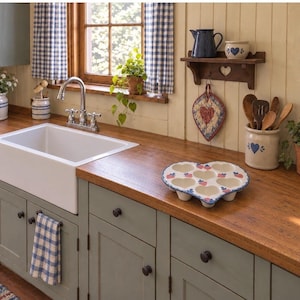 May include: A white ceramic heart-shaped muffin pan with a blue rim and small red heart patterns sits on a wooden countertop. The kitchen features a white farmhouse sink, a wooden shelf with a dark blue pitcher, and a ceramic crock holding wooden spoons.
