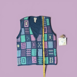 May include: A blue, patchwork-style crochet vest with a V-neck and button closure. The vest features various square and rectangular patterns in shades of blue, purple, and green. A yellow measuring tape is placed next to the vest, showing a length of approximately 28 inches.