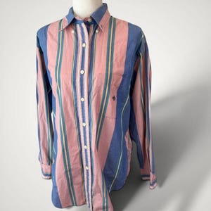 May include: A button-down shirt with vertical stripes in pink, blue, and yellow. The long-sleeved shirt has a button-down collar and a small logo on the chest. The shirt is displayed on a mannequin.