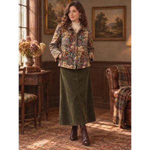 May include: A long-sleeved jacket featuring a dense, multi-colored floral tapestry pattern in shades of blue, orange, and cream, worn over a cream-colored mock neck top and a solid olive green ankle-length skirt with brown leather boots.