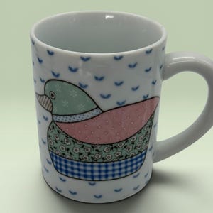 May include: White ceramic mug featuring a colorful duck design. The duck has a blue head, pink body, and a patterned wing. The mug has a white handle and is decorated with blue, stylized leaf shapes.