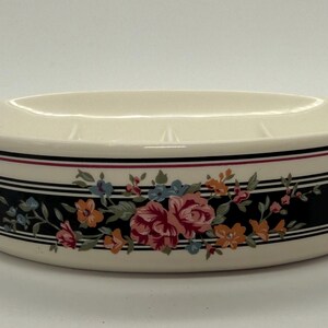 May include: Cream-colored ceramic soap dish with a floral design. The oval dish features a black band with a floral pattern of pink, orange, and blue flowers. The rim has black and red stripes.