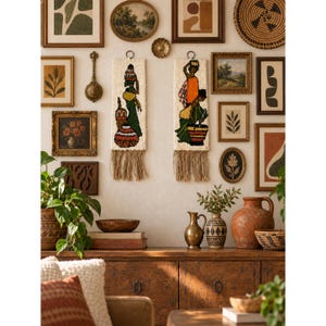 May include: Two rectangular off-white wall hangings featuring tufted figures carrying baskets and pots on their heads, finished with long fringe at the bottom. The figures are depicted in vibrant green, orange, and red tones against a neutral background.