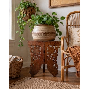 May include: A round ceramic planter with a tan and brown geometric pattern sits atop a dark brown wooden side table featuring intricate leaf carvings. The planter holds a lush green pothos plant, with another hanging in a woven basket nearby.