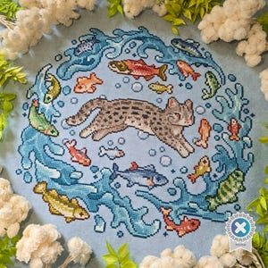 May include: A cross-stitch embroidery on light blue fabric featuring a central brown spotted cat surrounded by a circular pattern of various colorful fish and blue water splashes, with small white bubbles interspersed throughout the design.