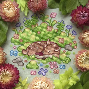 May include: A cross-stitch design featuring a sleeping fawn nestled in a bed of green foliage and colorful flowers. The fawn is brown with a white belly. The design is surrounded by dried flowers in shades of pink, peach, and burgundy.