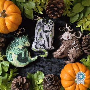 May include: Three embroidered keychains featuring pixelated creature designs: a green dragon, a grey gargoyle, and a brown horned beast. Each keychain includes a metal lobster clasp. The designs are crafted with detailed cross-stitch patterns.