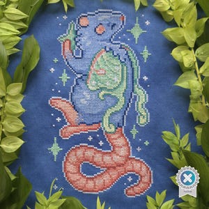 May include: Cross-stitch art featuring a blue creature with a rat-like head and a red tail, holding a green object. The design includes green star accents on a blue background. The artwork is surrounded by green leaves.