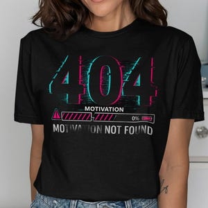 May include: Black t-shirt with a glitch effect design. The graphic reads "404 Motivation" in teal and pink, with a loading bar showing 0% and the text "Motivation Not Found".