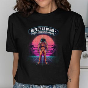 Puede incluir: Camiseta negra con un gráfico retro de un astronauta frente a una puesta de sol con palmeras. El texto "DEPLOY AT DAWN - PRODUCTION WAITS FOR NO ONE" está encima del astronauta. Un diseño único para los entusiastas del espacio.