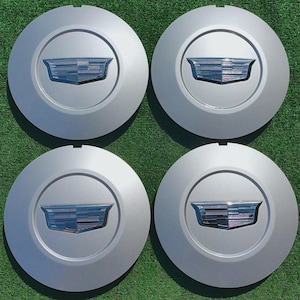 Puede incluir: Cuatro tapacubos plateados con el emblema de Cadillac en el centro. El emblema es un escudo estilizado con un diseño plateado y azul. Los tapacubos son circulares y tienen un acabado liso y mate.