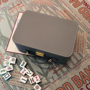 Puede incluir: Un juego de Mahjong gris con un estuche a juego, asa y cierre dorado. Las fichas son blancas con s&iacute;mbolos rojos, verdes y negros. El juego est&aacute; sobre un tablero de juego impreso con el texto "Bird Bam Mahjong Club."